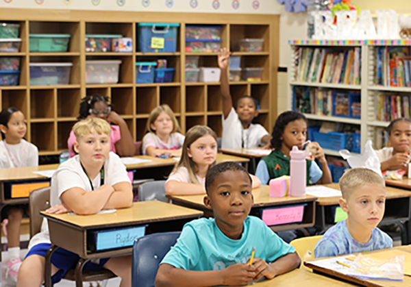 Elementary students sitting at desks with one student raising his hand.