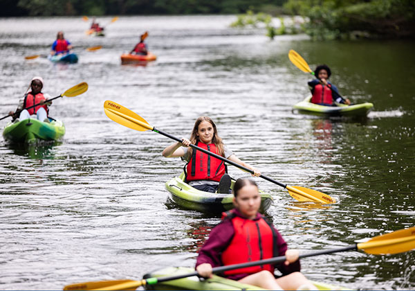 Students paddle kayaks in a line across a lake, wearing red life jackets and holding yellow paddles.
